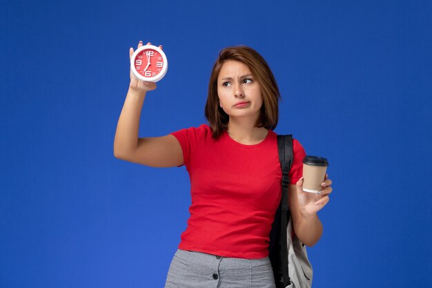 Vooraanzicht van vrouwelijke student in rood overhemd met klokken en koffie van de rugzakholding op de blauwe muur