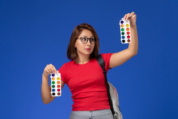 Vooraanzicht van vrouwelijke student in rood overhemd met rugzak die verf voor tekening op blauwe muur houdt