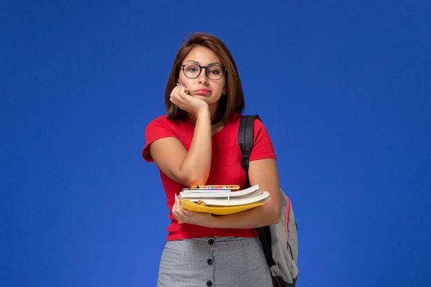 Vooraanzicht van vrouwelijke student in rood shirt met rugzak met boeken en bestanden op de lichtblauwe muur