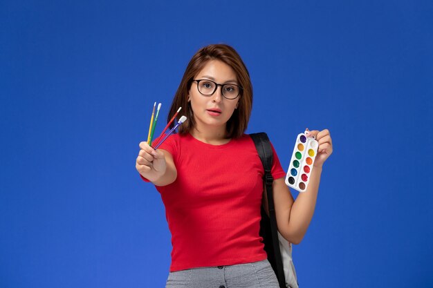 Vooraanzicht van vrouwelijke student in rood shirt met rugzak met verf voor tekening en kwastjes op de blauwe muur