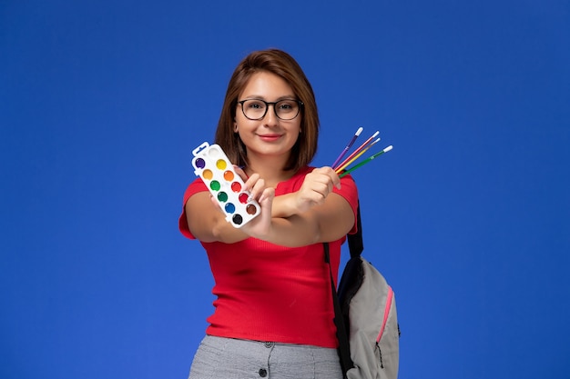 Vooraanzicht van vrouwelijke student in rood shirt met rugzak met verf voor tekening en kwastjes op de blauwe muur