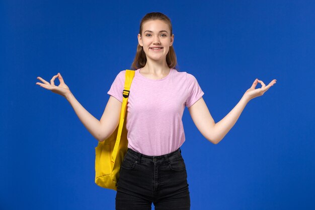 Vooraanzicht van vrouwelijke student in roze t-shirt met gele rugzak glimlachend en poseren op de blauwe muur