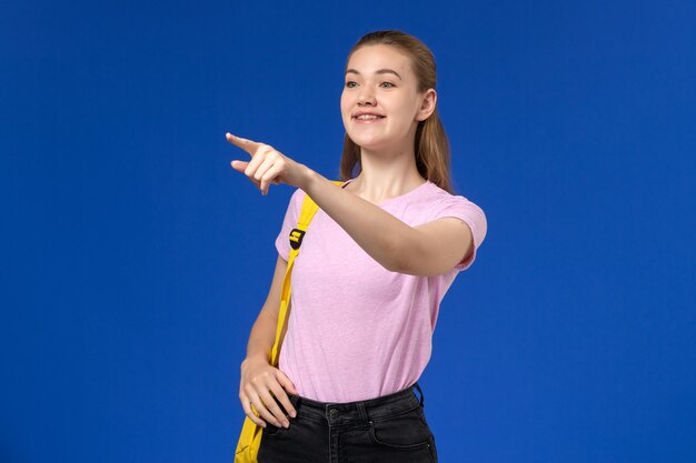 Vooraanzicht van vrouwelijke student in roze t-shirt met gele rugzak glimlachend op de blauwe muur