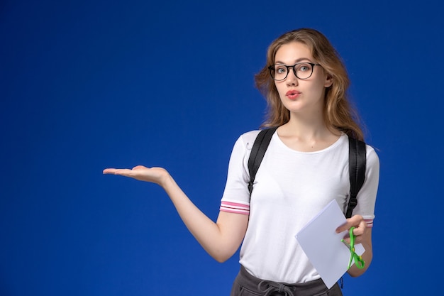 Vooraanzicht van vrouwelijke student in wit overhemd die rugzak draagt en document en schaar op de blauwe muur houdt