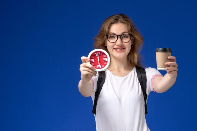 Vooraanzicht van vrouwelijke student in wit overhemd die rugzak draagt en klokken en koffie houdt die op de blauwe muur glimlachen