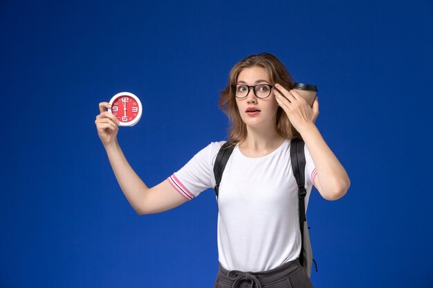 Vooraanzicht van vrouwelijke student in wit overhemd die rugzak draagt en klokken en koffie op de blauwe muur houdt