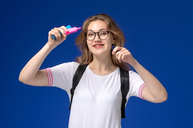 Vooraanzicht van vrouwelijke student in wit overhemd die rugzak draagt en viltstiften op de blauwe muur houdt