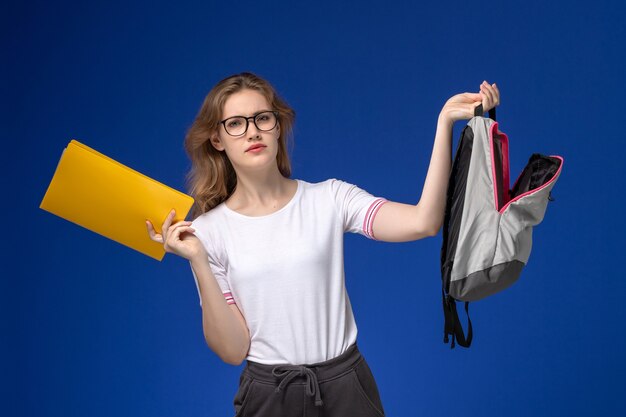 Vooraanzicht van vrouwelijke student in wit overhemd met rugzak en gele bestanden op de blauwe muur