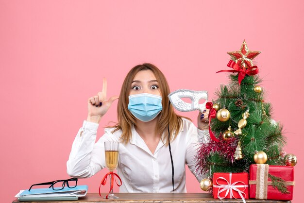 Vooraanzicht van vrouwelijke werknemer in masker zittend achter haar tafel met kerstcadeautjes op roze