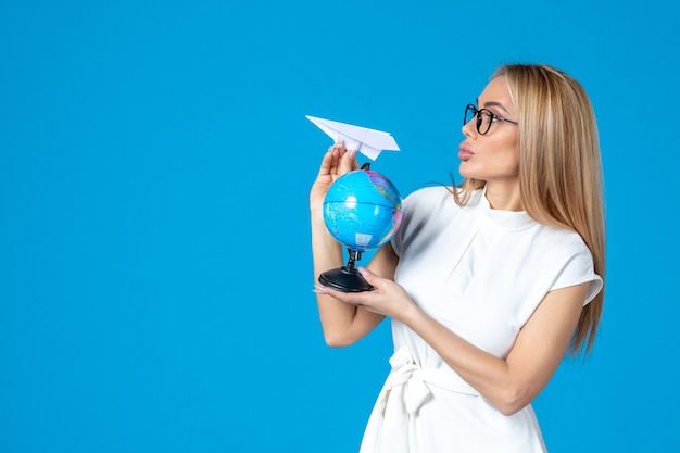 Vooraanzicht van vrouwelijke werknemer in witte jurk met earth globe en papieren vliegtuigje op blauwe muur
