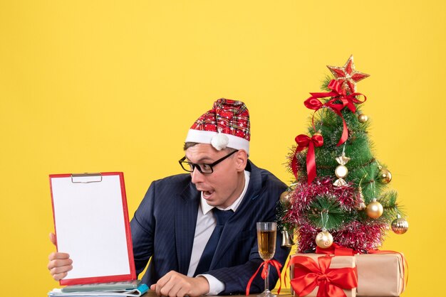 Vooraanzicht van zakenman kijken naar klembord zittend aan de tafel in de buurt van kerstboom en presenteert op geel