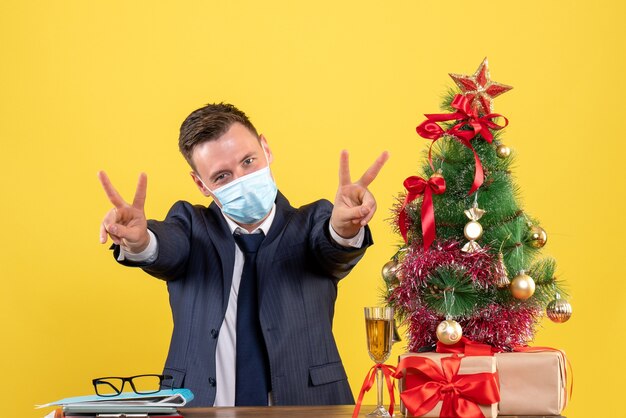 Vooraanzicht van zakenman maken overwinning teken zittend aan de tafel in de buurt van kerstboom en presenteert op geel