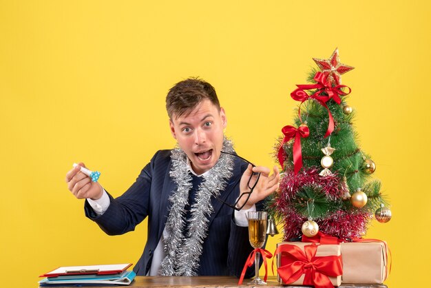 Vooraanzicht van zakenman met noisemaker zittend aan de tafel in de buurt van kerstboom en presenteert op geel