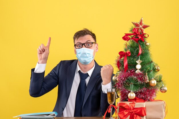 Vooraanzicht van zakenman wijzend met vinger omhoog zitten aan de tafel in de buurt van kerstboom en presenteert op geel