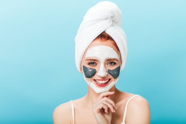 Vooraanzicht van zalige blanke vrouw met gezichtsmasker. Studio shot van aangenaam meisje met handdoek op hoofd poseren op blauwe achtergrond.