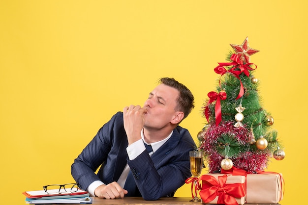 Vooraanzicht van zalige man kus gebaar zittend aan de tafel in de buurt van kerstboom en cadeautjes op gele muur