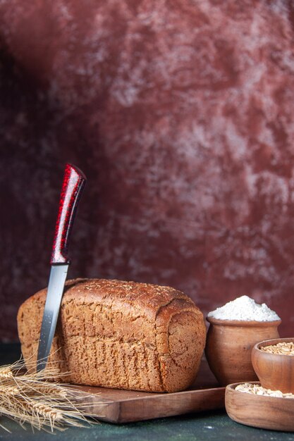 Vooraanzicht van zwart brood sneetjes meel in een kom op een houten bord en mes spikes rauwe havermout tarwe op gemengde kleuren noodlijdende achtergrond