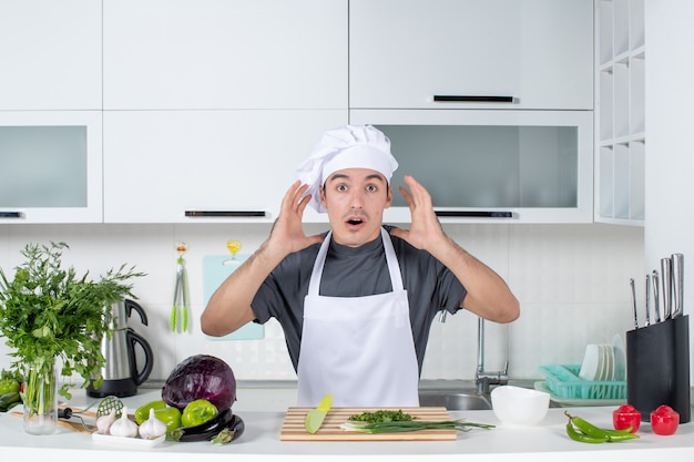 Vooraanzicht verbaasde mannelijke chef-kok in uniform die achter de keukentafel staat