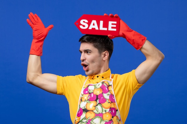 Gratis foto vooraanzicht verbaasde mannelijke huishoudster in geel t-shirt met verkoopbord boven zijn hoofd op blauwe ruimte