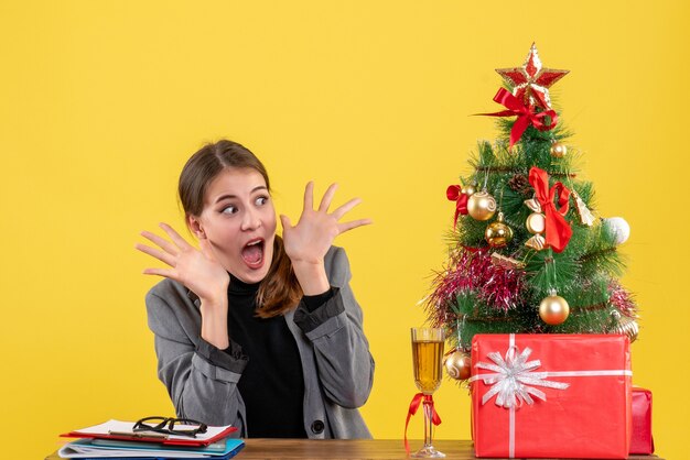 Vooraanzicht verrast schattig meisje zit aan de balie met geopende handen kerstboom en geschenken cocktail