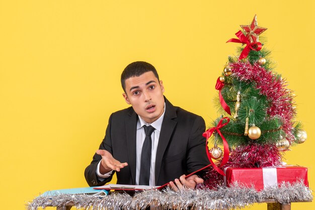 Vooraanzicht verwarde man in pak zittend aan de tafel met document kerstboom en geschenken