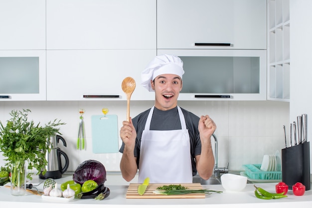 Vooraanzicht vrolijke mannelijke chef-kok in uniform met houten lepel achter keukentafel