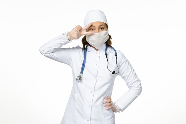 Vooraanzicht vrouwelijke arts in wit medisch pak en met masker wegens coronavirus op witte muurvirusziekte pandemische covid