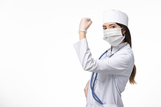 Vooraanzicht vrouwelijke arts in wit medisch pak met een masker vanwege coronavirus buigen op witte muur gezondheid ziekte pandemie covid-
