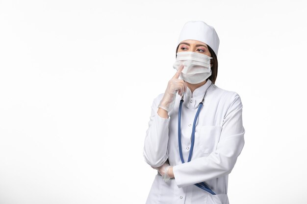 Vooraanzicht vrouwelijke arts in wit medisch pak met een masker vanwege coronavirus diep na te denken over witte muur gezondheid ziekte pandemie covid-