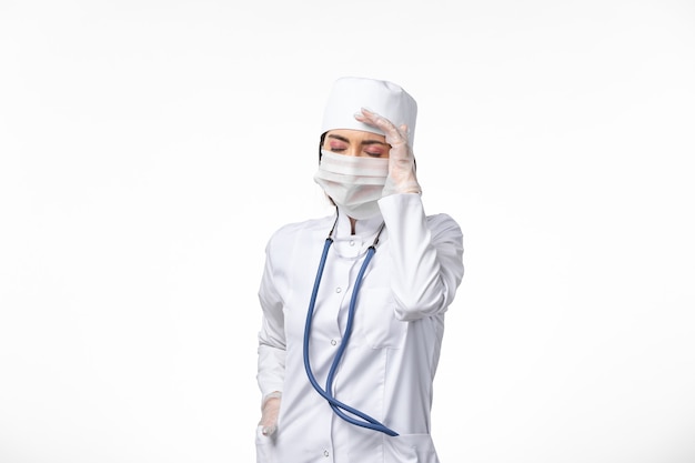 Vooraanzicht vrouwelijke arts in wit medisch pak met een masker vanwege coronavirus met hoofdpijn op witte muur ziekte geneeskunde virus pandemie covid-