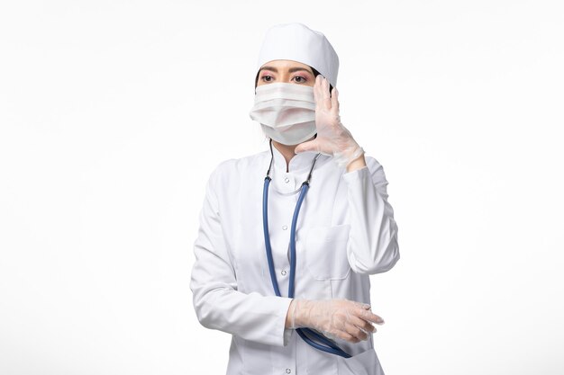 Vooraanzicht vrouwelijke arts in wit medisch pak met een masker vanwege coronavirus op witte muur ziekte geneeskunde virus pandemie covid-
