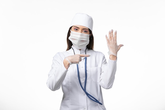 Vooraanzicht vrouwelijke arts in wit steriel medisch pak met masker vanwege covid- toont haar hand op het witte bureau ziekte covid- pandemische virusziekte