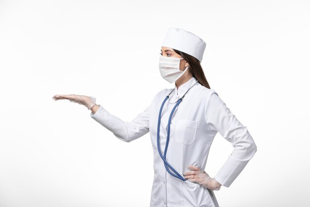 Vooraanzicht vrouwelijke arts in wit steriel medisch pak met masker vanwege covid- toont haar hand op witte muurziekte covid- pandemische virusziekte