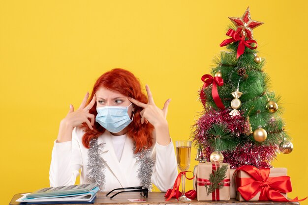 Vooraanzicht vrouwelijke arts rond kerstboom en cadeautjes zitten in masker