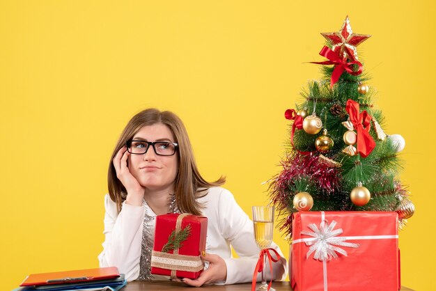 Vooraanzicht vrouwelijke arts zit tafel met cadeautjes en boom op gele achtergrond met kerstboom en geschenkdozen