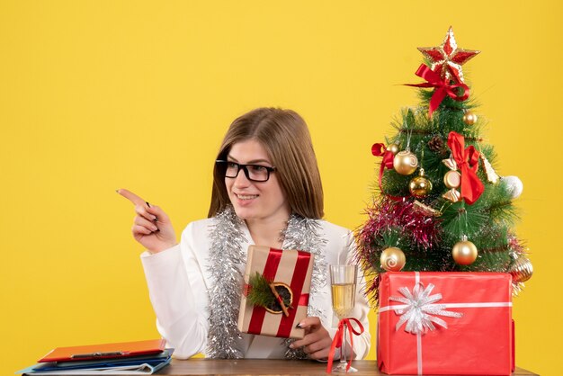 Vooraanzicht vrouwelijke arts zit voor haar tafel bedrijf aanwezig op geel bureau met kerstboom en geschenkdozen