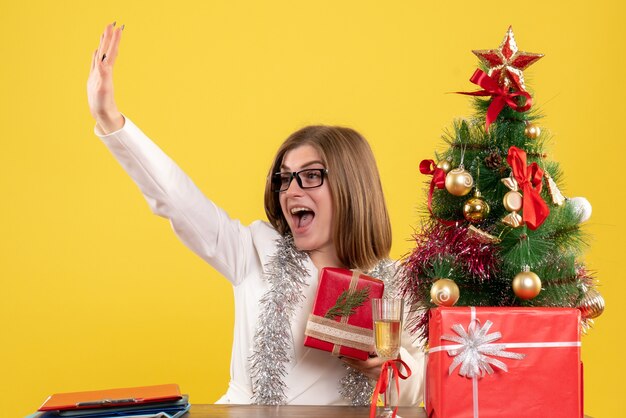 Vooraanzicht vrouwelijke arts zit voor haar tafel bedrijf aanwezig op gele achtergrond met kerstboom en geschenkdozen