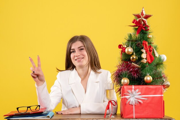 Vooraanzicht vrouwelijke arts zit voor haar tafel op gele achtergrond met kerstboom en geschenkdozen