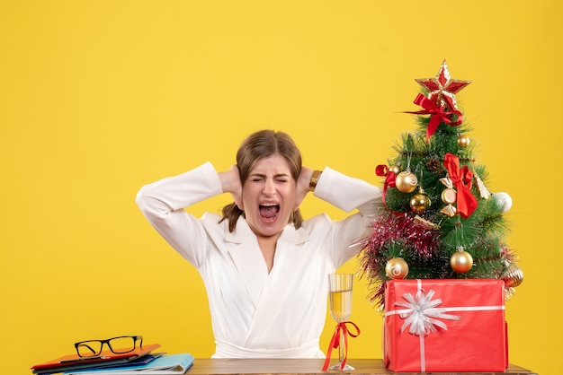 Vooraanzicht vrouwelijke arts zit voor haar tafel op gele achtergrond met kerstboom en geschenkdozen