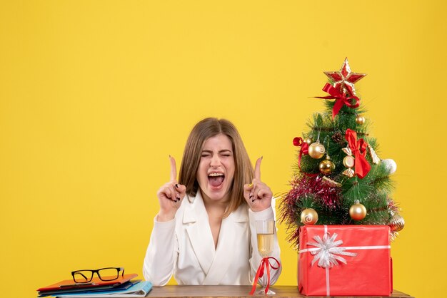 Vooraanzicht vrouwelijke arts zit voor haar tafel op gele achtergrond met kerstboom en geschenkdozen