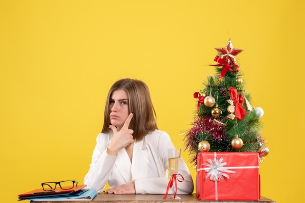Vooraanzicht vrouwelijke arts zit voor haar tafel op gele achtergrond met kerstboom en geschenkdozen