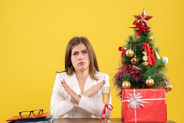 Vooraanzicht vrouwelijke arts zit voor haar tafel op gele achtergrond met kerstboom en geschenkdozen