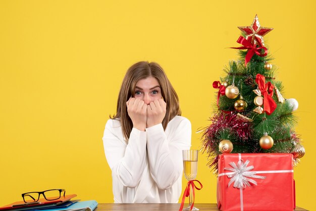 Vooraanzicht vrouwelijke arts zit voor haar tafel op gele achtergrond met kerstboom en geschenkdozen