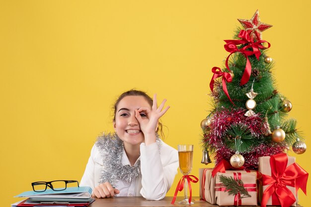 Vooraanzicht vrouwelijke arts zittend achter haar tafel lachend op gele achtergrond met kerstboom en geschenkdozen