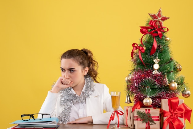 Vooraanzicht vrouwelijke arts zittend achter haar tafel met gestrest gezicht op gele achtergrond met kerstboom en geschenkdozen