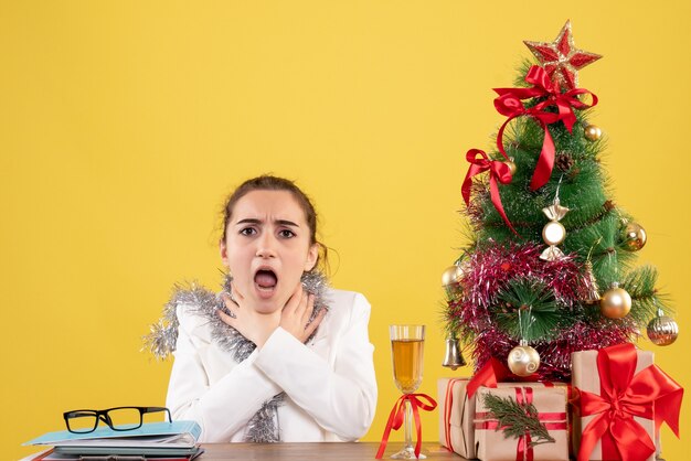 Vooraanzicht vrouwelijke arts zittend achter haar tafel met keelproblemen op gele achtergrond met kerstboom en geschenkdozen