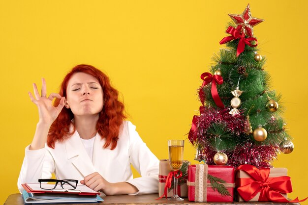 Gratis foto vooraanzicht vrouwelijke arts zittend achter haar tafel met kerstcadeautjes en boom op geel