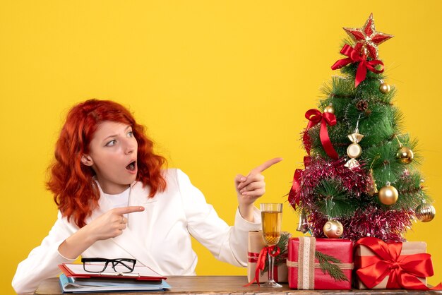 Vooraanzicht vrouwelijke arts zittend achter haar tafel met kerstcadeautjes en boom op gele achtergrond