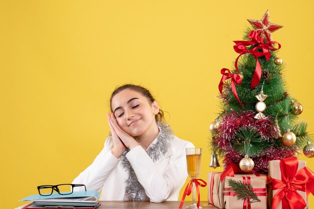 Vooraanzicht vrouwelijke arts zittend achter haar tafel met opgetogen gezicht op gele achtergrond met kerstboom en geschenkdozen