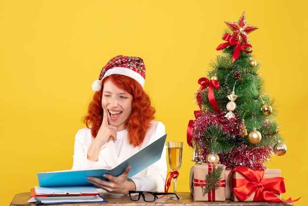 Vooraanzicht vrouwelijke arts zittend achter tafel documenten op de gele achtergrond met kerstboom en geschenkdozen te lezen